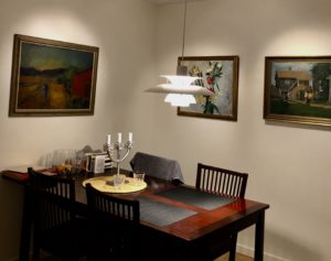 Matplats i ett seniorboende i Stockholm med ett mörkt träbord, fyra stolar, och en modern taklampa som hänger ovanför. Väggarna pryds av målningar och en kandelaber står på bordet. En referenskund har fått professionell belysningskonsultation för att förbättra ljusmiljön.