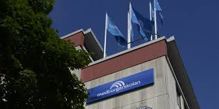 Fasad av Medborgarskolan byggnad med deras blåa flaggor vajande på taket mot en klarblå himmel. 2700kelvin studerade diplomutbildningen Inredning rum och form hos Medborgarskolan.