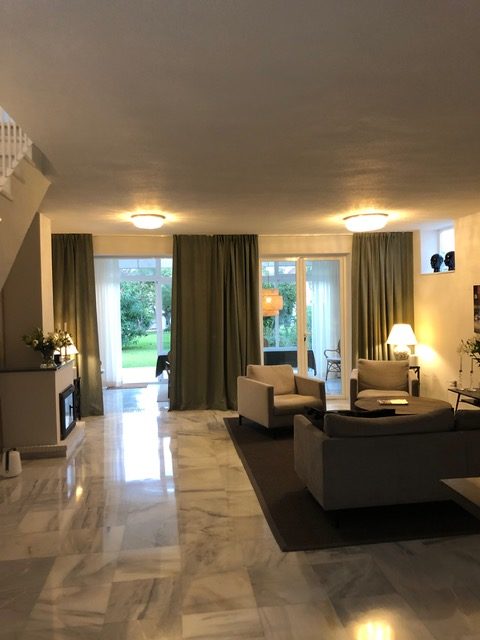 Rymligt vardagsrum i ett spanskt hem med marmorgolv, stora fönster med gröna gardiner, och flera bekväma sittplatser. En referenskund i Spanien har fått online belysningskonsultation för att skapa en varm och välbelyst atmosfär.