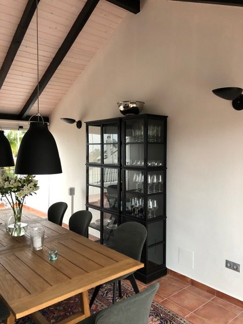 Matplats i ett spanskt hem med lutande tak, svarta hängande lampor över ett träbord, och en vitrinskåp fylld med glas. En referenskund i Spanien har fått online belysningskonsultation för att optimera ljuset i sitt hem.